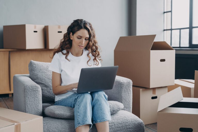 Focused hispanic girl works at laptop searching ideas for renovation, choosing furniture, shopping interior decor for new house online, sitting on armchair among card boxes. Moving, relocation concept